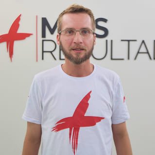 Pedro Roberts, coordenador de tráfego pago, usando camiseta branca com estampa vermelha, em frente ao logotipo da Agência Mais Resultado.